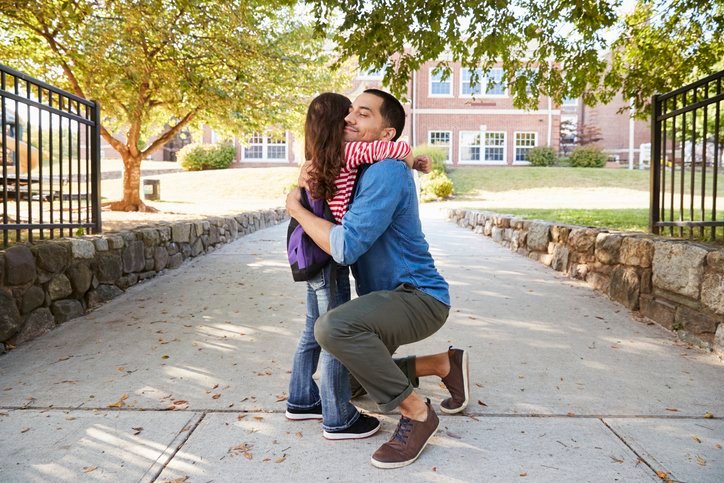 Parent kneeling to hug a child goodbye outside a school entrance