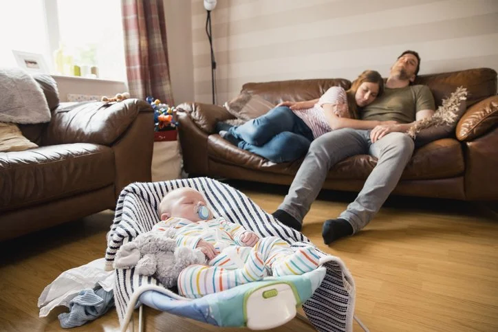 Exhausted parents resting on the couch while their baby sleeps nearby
