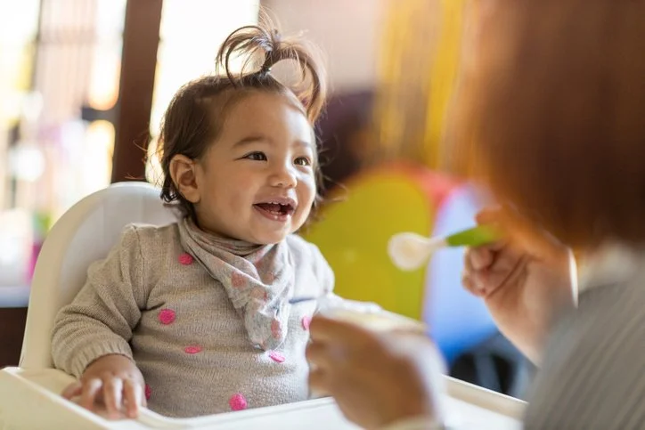 Toddler smiling in a high chair while being fed by a parent
