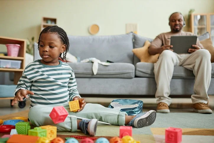 Toddler stacking colorful blocks on the floor while a parent works nearby