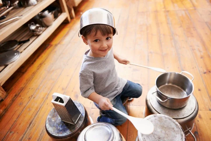 Child playfully drumming on pots and pans with a pot on their head