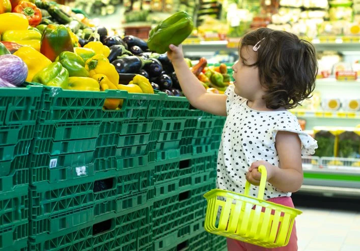 Toddler reaching for produce while holding a small shopping basket
