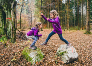 Two children playfully hopping between large rocks in a leaf-covered forest