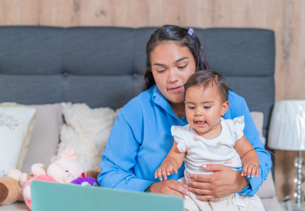 Parent sitting on a bed with a baby, looking at a laptop screen
