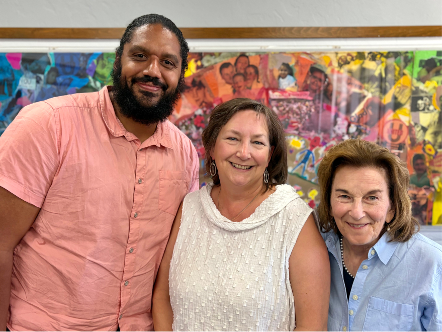 Three smiling adults standing together in front of a colorful mural